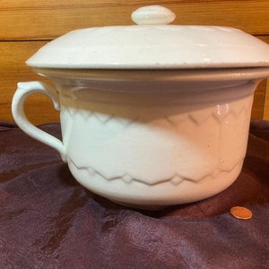 Puede incluir: Olla de cerámica blanca con tapa y asa. La olla tiene un diseño decorativo alrededor de la parte inferior.