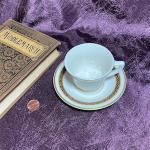 May include: A white teacup and saucer set with gold and black trim. The saucer and cup are on a purple fabric surface next to a book titled "Middlemarch" and a penny.