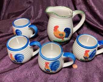 Puede incluir: Conjunto de cuatro tazas de cerámica blanca con diseños de gallo azul y rojo y una jarra de cerámica blanca a juego con un borde verde y un diseño de gallo.