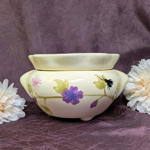 May include: A pale yellow ceramic planter with a scalloped rim and three small feet. The exterior features a hand-painted botanical design with green leaves, purple and pink flowers, and a small black insect, creating a delicate floral motif.