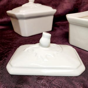 May include: White porcelain lidded rectangular dish with a decorative finial on the lid. The lid is off, revealing the interior. Two other similar dishes are in the background, all on a dark purple surface.