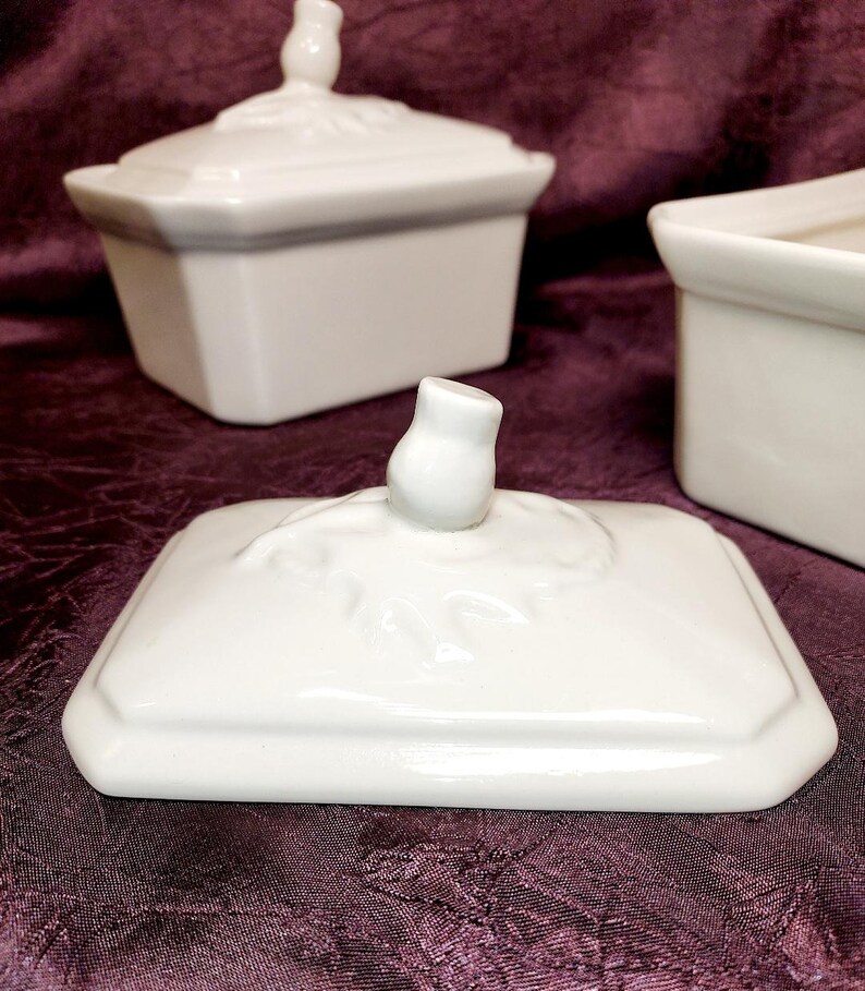 May include: White porcelain lidded rectangular dish with a decorative finial on the lid. The lid is off, revealing the interior. Two other similar dishes are in the background, all on a dark purple surface.