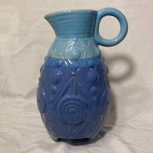 May include: A blue ceramic pitcher with a textured design and a handle. The pitcher has a light blue top and a darker blue bottom.