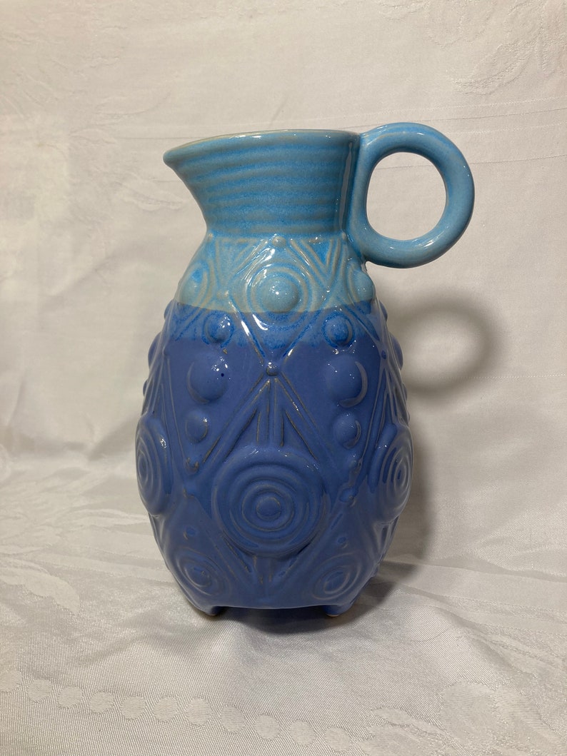 May include: A blue ceramic pitcher with a textured design and a handle. The pitcher has a light blue top and a darker blue bottom.