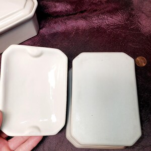 May include: A collection of white ceramic ashtrays and a matching lid. The ashtrays are rectangular with a small notch on one side. A penny is next to the stack of ashtrays for size comparison. The background is a dark purple fabric.