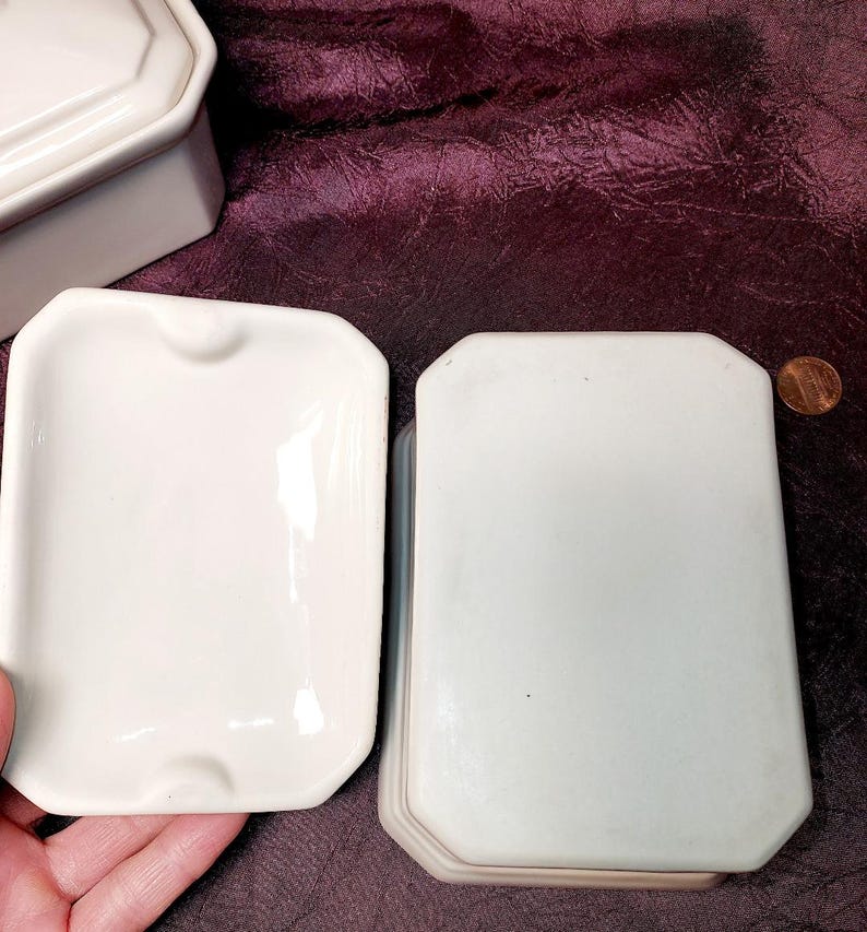 May include: A collection of white ceramic ashtrays and a matching lid. The ashtrays are rectangular with a small notch on one side. A penny is next to the stack of ashtrays for size comparison. The background is a dark purple fabric.