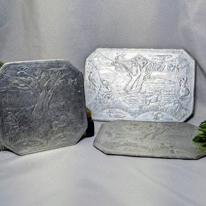 May include: Three silver-toned metal trays with embossed landscape scenes featuring trees, animals, and figures. The trays have octagonal or rectangular shapes with beveled edges and detailed relief carvings depicting outdoor nature settings.