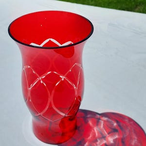 May include: A bright red glass vase with a black rim and an etched design. The vase's shadow is visible on the white surface. The vase is a decorative item.