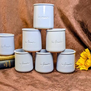 May include: A collection of six light blue ceramic yogurt pots stacked in a pyramid formation. Each pot features a smooth, cylindrical shape with a slightly flared rim and the embossed text la fermière on the front surface of the exterior.