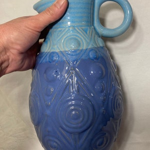May include: A blue ceramic pitcher with a textured design of circles and triangles. The pitcher has a handle and three feet.