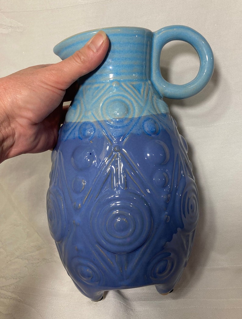 May include: A blue ceramic pitcher with a textured design of circles and triangles. The pitcher has a handle and three feet.
