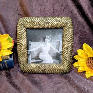 May include: A square picture frame with a textured, gold-colored snakeskin pattern border. The frame contains a black and white photograph of a person wearing a light-colored dress, seated on a bench with their arms resting along the backrest.