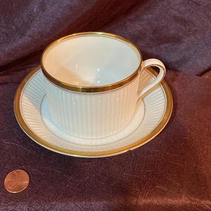 Puede incluir: Una taza de té blanca con un borde dorado y un platillo a juego. La taza de té tiene líneas verticales y el platillo tiene un borde dorado.
