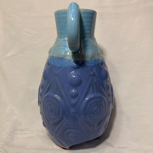 May include: A blue ceramic pitcher with a textured design and a handle. The pitcher has a light blue top and a darker blue bottom.
