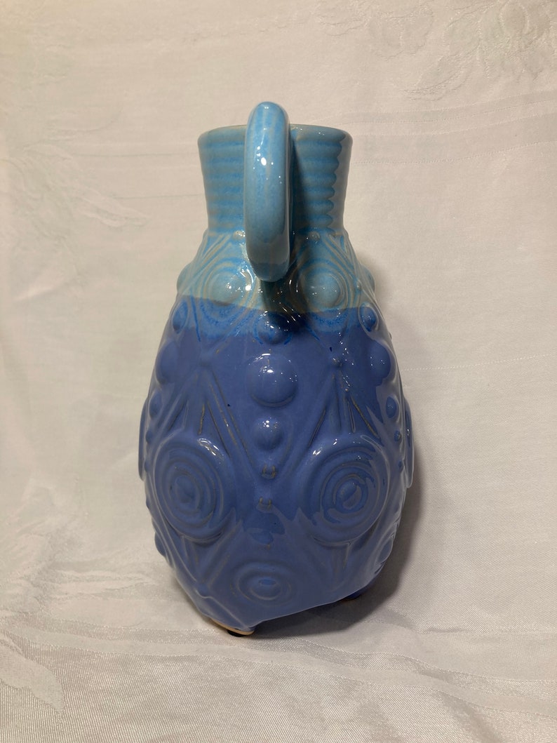 May include: A blue ceramic pitcher with a textured design and a handle. The pitcher has a light blue top and a darker blue bottom.
