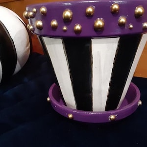 Op de afbeelding: Paarse en wit gestreepte plantenpot met een gouden rand met studs. De pot heeft een bijpassende paarse schotel met gouden studs.