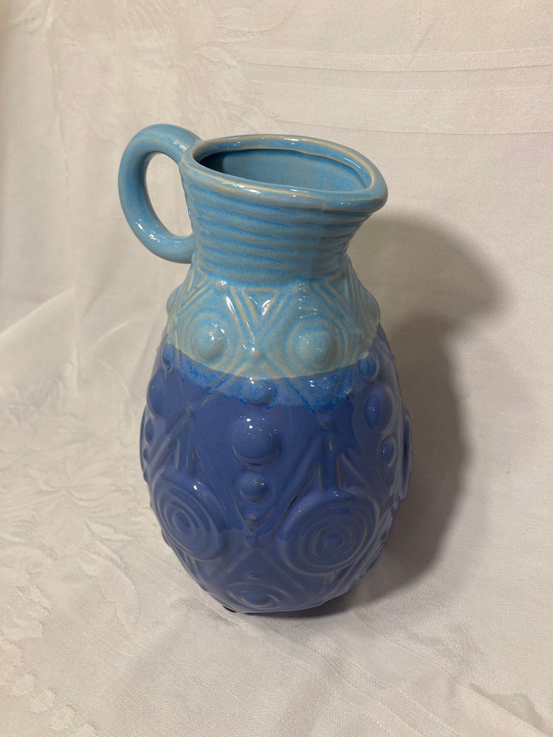 May include: A blue ceramic pitcher with a textured design. The pitcher has a light blue top and a darker blue bottom. The pitcher has a handle and a round body.