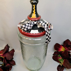 Puede incluir: Un frasco de vidrio transparente con superficie texturizada y tapa decorativa. La tapa está pintada de negro con puntos blancos, un borde rojo y una cinta a cuadros blancos y negros atada en la base. Un remate negro y rojo corona la tapa.