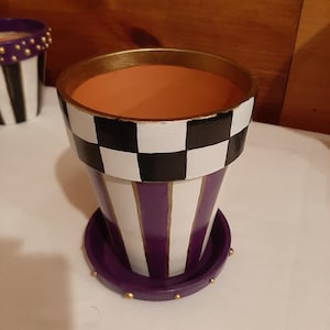 May include: A purple and white striped planter with a black and white checkered top. The planter has a gold rim and a purple saucer with gold dots.