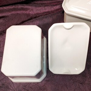 May include: White ceramic butter dish with a lid and a separate dish. The dish has a rectangular shape with rounded corners. The lid is also rectangular with a slightly raised design. The set is on a dark purple surface.