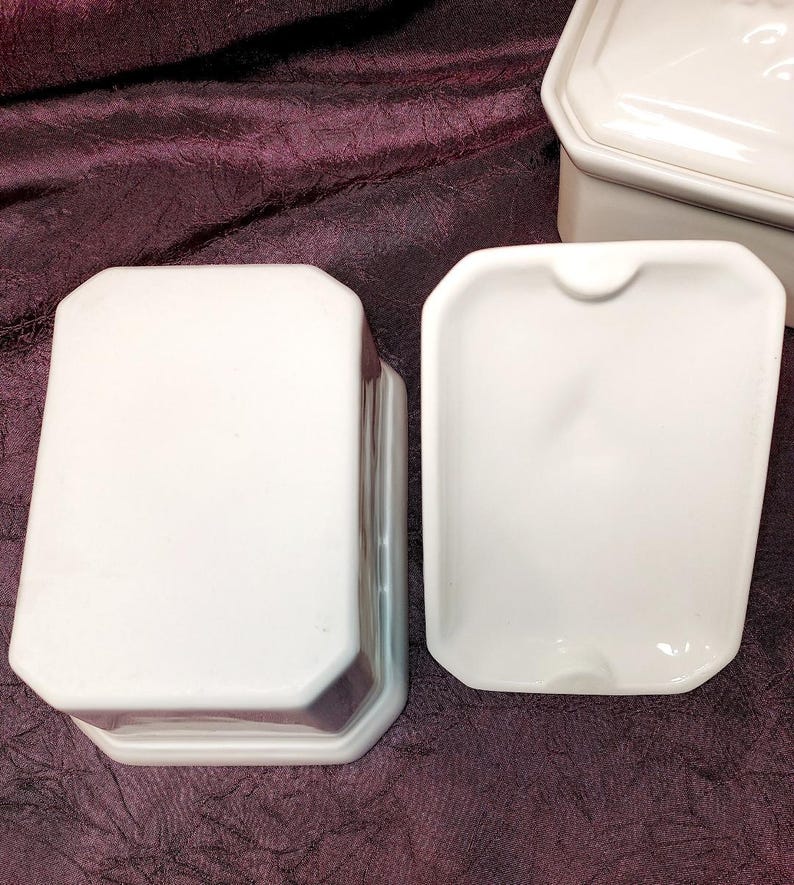 May include: White ceramic butter dish with a lid and a separate dish. The dish has a rectangular shape with rounded corners. The lid is also rectangular with a slightly raised design. The set is on a dark purple surface.