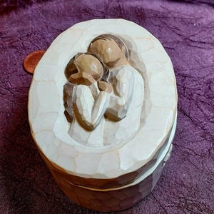 Könnte beinhalten: Kleine, ovale Schmuckschatulle in Off-White mit einem geschnitzten Relief von zwei sich umarmenden Figuren. Die Figuren sind in einem einfachen, rustikalen Stil gehalten. Die Box hat ein strukturiertes, geschnitztes Aussehen.