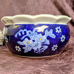 May include: A round ceramic bowl with a scalloped rim, featuring a deep cobalt blue glaze decorated with light blue floral patterns and small white flowers with yellow centers. The interior and the top edge of the bowl are finished in white.