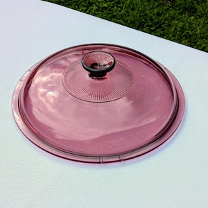 May include: A round, translucent pink glass lid featuring a smooth, domed surface and a central knob. The top of the lid displays a radial, sunburst-like pattern of fine, etched lines radiating outward from the base of the small, circular handle.
