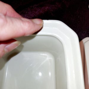 May include: Close-up of a white rectangular ceramic dish with rounded corners. The dish has a glossy finish and is partially visible, with a finger touching the edge. The background is a dark, textured fabric.