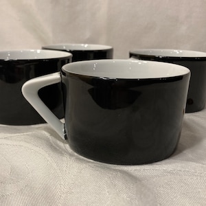 May include: Four black and white ceramic mugs with white interiors. The mugs have a glossy finish and are arranged in a row on a white surface.
