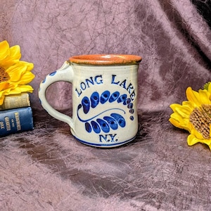 May include: A ceramic mug with a speckled cream glaze, featuring a brown rim and a blue hand-painted leaf design. The text "LONG LAKE N.Y." is inscribed in blue on the front. The mug has a curved handle and a sturdy base, showcasing a handcrafted aesthetic.