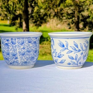May include: Two white ceramic bowls with blue painted designs. The left bowl features a dense pattern of small flowers and swirling vines. The right bowl displays a sparse pattern of elongated leaves and small berries arranged on thin stems.