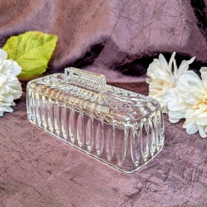 May include: A rectangular clear glass butter dish cover featuring a vertical ribbed pattern along the sides and a smooth, raised rectangular handle on top. The glass is transparent with a reflective surface, showcasing a structured, geometric design.