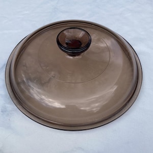Op de afbeelding: Een bruine glazen deksel voor een pot of pan. De deksel heeft een ronde knop bovenop.