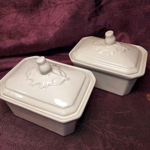 May include: Two off-white rectangular ceramic dishes with lids. Each lid features a raised floral design and a small knob. The dishes are on a dark purple surface. The dishes are likely for serving or storage.