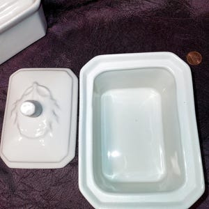 May include: White ceramic butter dish with a lid, featuring a rectangular shape with beveled edges. The lid has a decorative knob and embossed leaf design. The dish is open, revealing its interior. A penny is visible in the background.