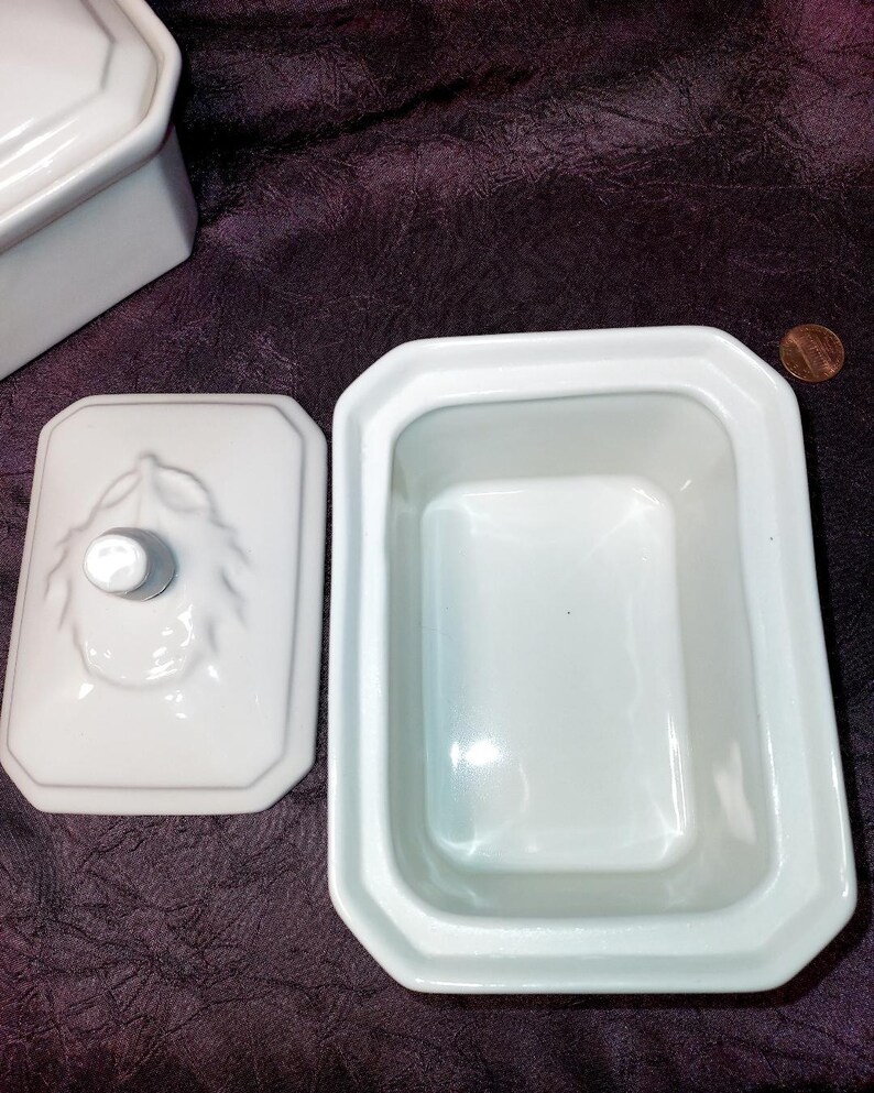 May include: White ceramic butter dish with a lid, featuring a rectangular shape with beveled edges. The lid has a decorative knob and embossed leaf design. The dish is open, revealing its interior. A penny is visible in the background.