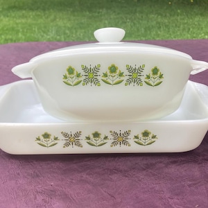 Könnte beinhalten: Eine weiße Auflaufform mit grünem Blumenmuster. Die Form ist aus Pyrex und hat einen Deckel. Die Form steht auf einer violetten Oberfläche.