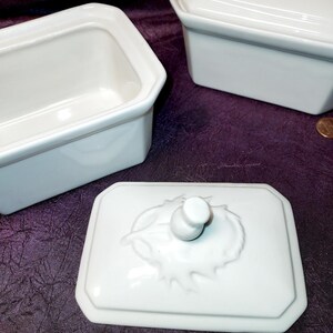 May include: White rectangular ceramic dish with a lid featuring a leaf design and a small knob. The dish has a simple, elegant design. Two other similar dishes are also visible.