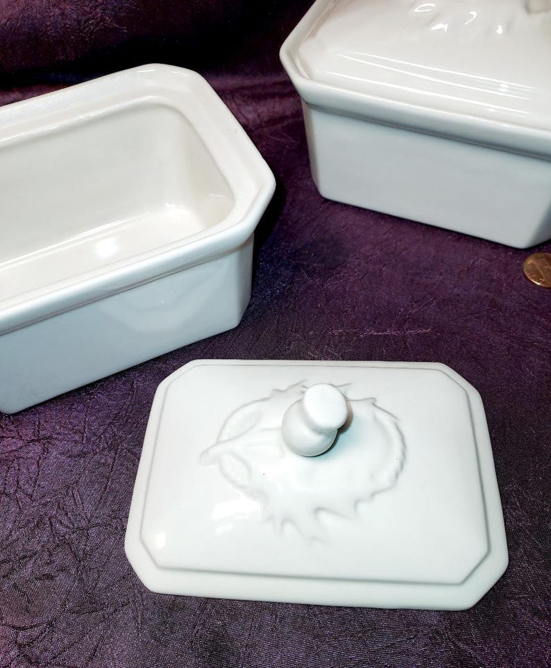 May include: White rectangular ceramic dish with a lid featuring a leaf design and a small knob. The dish has a simple, elegant design. Two other similar dishes are also visible.