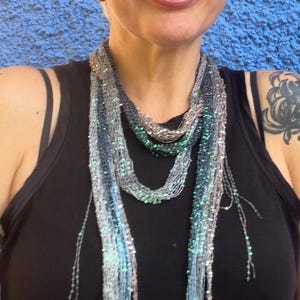 May include: A beaded scarf in shades of grey, blue, and green. The scarf is worn over a black vest top. The background is a textured blue wall. The scarf has a delicate, flowing design.