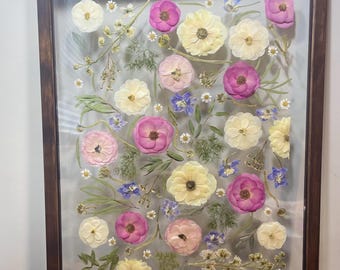 May include: A framed pressed flower art piece with a variety of flowers in shades of pink, white, and blue. The flowers are arranged in a random pattern and are pressed between two pieces of glass.