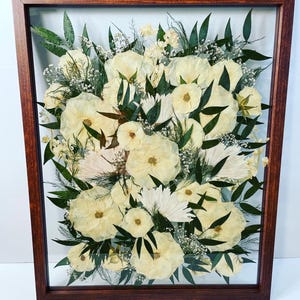 May include: A framed floral arrangement featuring cream-colored flowers, green leaves, and delicate white accents. The flowers are arranged in a dense, layered composition within a dark brown wooden frame, creating a natural, elegant display.