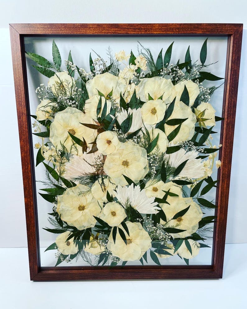 May include: A framed floral arrangement featuring cream-colored flowers, green leaves, and delicate white accents. The flowers are arranged in a dense, layered composition within a dark brown wooden frame, creating a natural, elegant display.