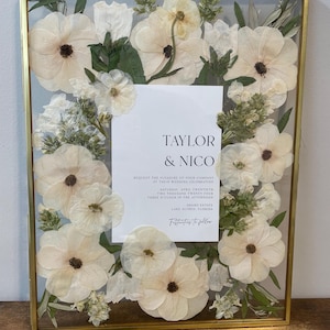 May include: A gold-framed glass box with dried white flowers and greenery. The box contains a wedding invitation for Taylor & Nico, with the date and location of the wedding.