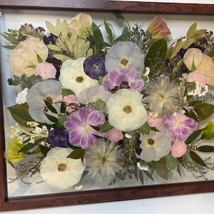 May include: A framed floral arrangement featuring a variety of dried flowers in shades of white, pink, purple, and yellow. The flowers are arranged in a dense, layered composition, encased in a wooden frame.