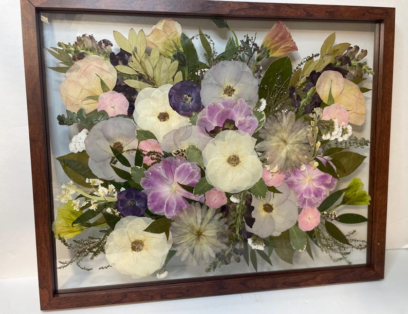 May include: A framed floral arrangement featuring a variety of dried flowers in shades of white, pink, purple, and yellow. The flowers are arranged in a dense, layered composition, encased in a wooden frame.