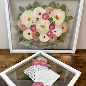 May include: Two white framed floral arrangements. The larger frame contains a bouquet of white, pink, and purple flowers with green leaves. The smaller frame holds a wedding invitation with floral accents and additional flowers.