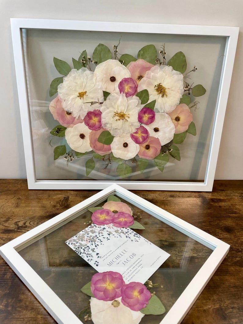 May include: Two white framed floral arrangements. The larger frame contains a bouquet of white, pink, and purple flowers with green leaves. The smaller frame holds a wedding invitation with floral accents and additional flowers.