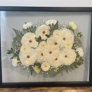 May include: A framed floral arrangement featuring cream-colored flowers with brown centers, surrounded by green foliage and baby's breath. The arrangement is encased in a black rectangular frame with a glass front.